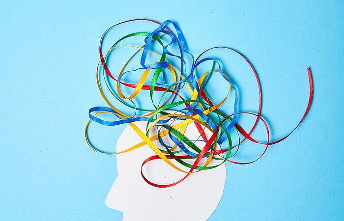 Silhouette eines menschlichen Kopfes in Weiß vor blauem Hintergrund, aus dem bunte Bänder in Rot, Blau, Grün und Gelb herauswirbeln, symbolisch für Gedanken, Kreativität und das Chaos im Kopf, passend zu ADS und ADHS.