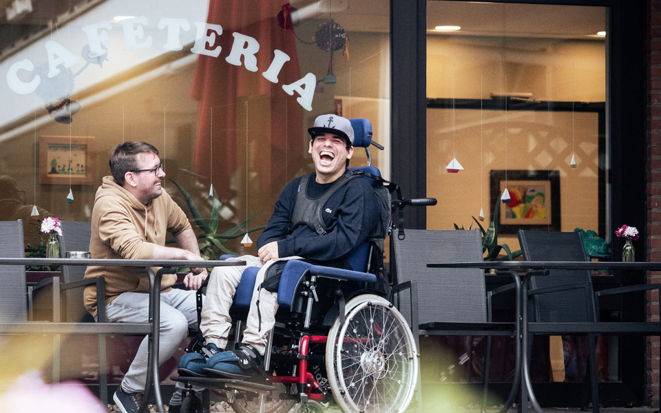 Zwei lachende Männer, einer im Rollstuhl, unterhalten sich auf der Terrasse der Cafeteria des Senator-Neumann-Hauses des Sozialkontors.