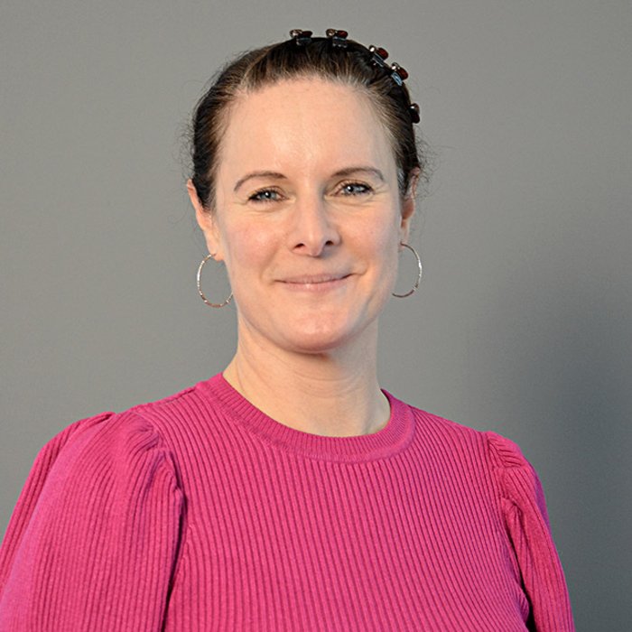 Anja Schubring-Wasiee, eine Frau mit zurückgebundenem braunen Haar, lächelt freundlich in die Kamera. Sie trägt ein pinkfarbenes Oberteil mit feinen Falten. Der Hintergrund ist neutral grau gestaltet.