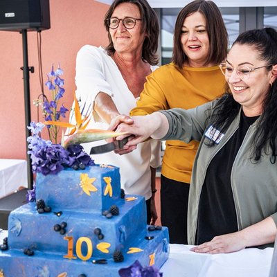 Drei Frauen (darunter Katharina Fegebank, Hamburgs Zweite Bürgermeisterin und Ina Terragnolo, die Leiterin des Haus Beerboom sowie eine weitere Mitarbeitende des Haus Beerboom) stehen gemeinsam an einem Tisch unter einem Pavillon und schneiden eine große, mehrstöckige Torte an. Die Torte ist blau mit bunten Zahlen und Symbolen dekoriert, obenauf blauer und lilafarbener Blumenschmuck. Im Hintergrund sind ein Gebäude und etwas Grün zu erkennen.