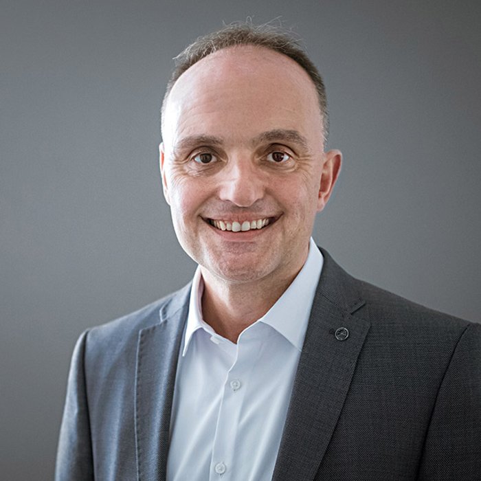 Michael Wild, ein Mann mit kurzem, grauem Haar und Brille, lächelt freundlich in die Kamera. Er trägt ein gemustertes Sakko und ein weißes Hemd. Der Hintergrund ist dunkelgrau gestaltet.