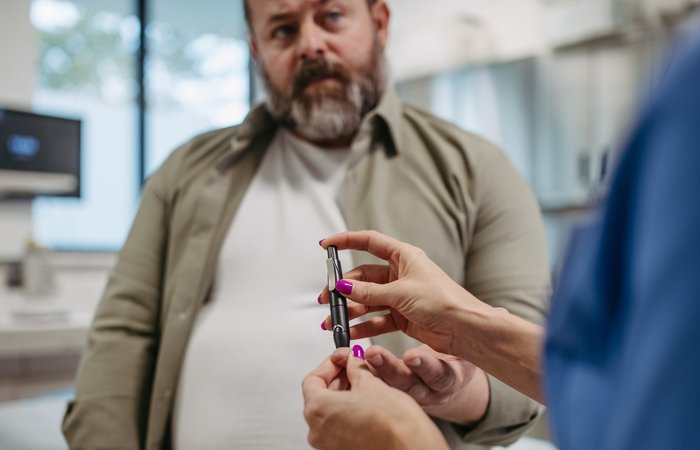 Mittelalter Mann mit Übergewicht, bei dem eine medizinische Fachkraft Blut aus dem Finger für einen Blutzucker-Test abnimmt.