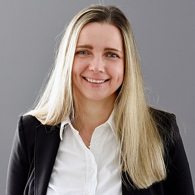 Karin Conradt, eine Frau mit langem, glatten, blonden Haar, lächelt freundlich in die Kamera. Sie trägt ein weißes Hemd unter einem schwarzen Blazer. Der Hintergrund ist neutral grau gestaltet.