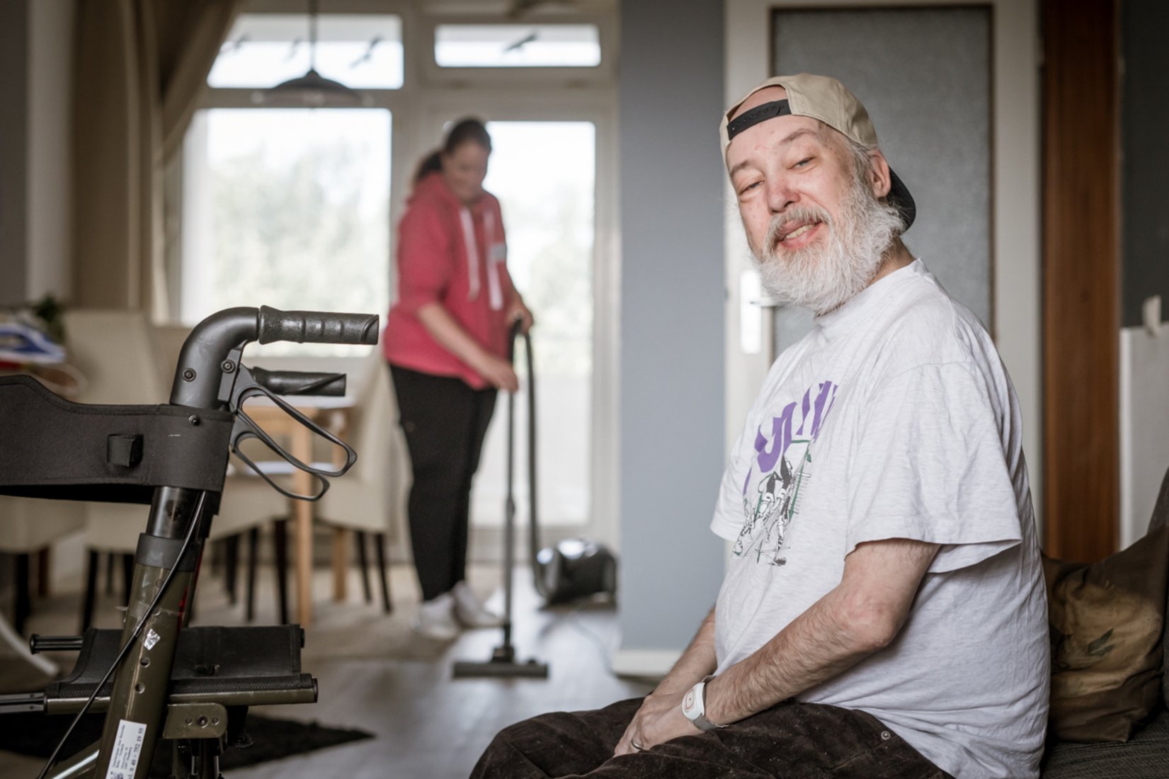 Ein älterer Mann mit grauem Bart, weißem T-Shirt und rückwärts getragener Kappe sitzt in einem Wohnzimmer und schaut freundlich in die Kamera. Im Hintergrund saugt eine Assistenzkraft den Boden. Ein Rollator steht neben dem Mann.