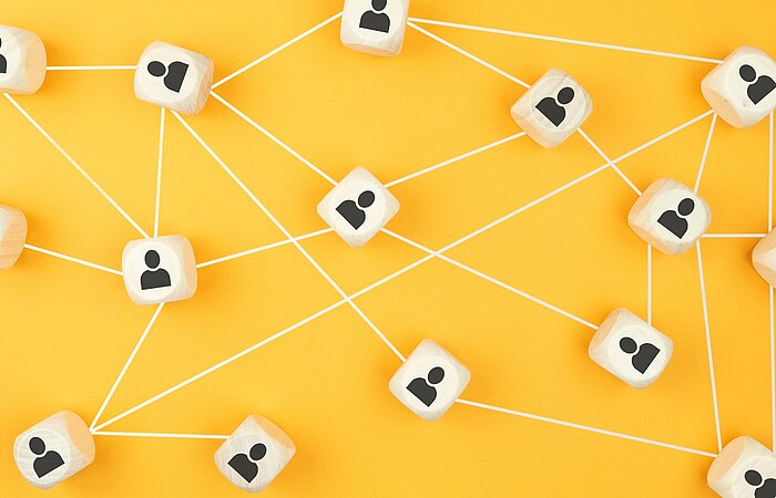 Holzwürfel mit Personen-Symbolen sind über weiße Linien auf gelbem Hintergrund miteinander verbunden und visualisieren ein Netzwerk aus Beziehungen und Kooperationen.