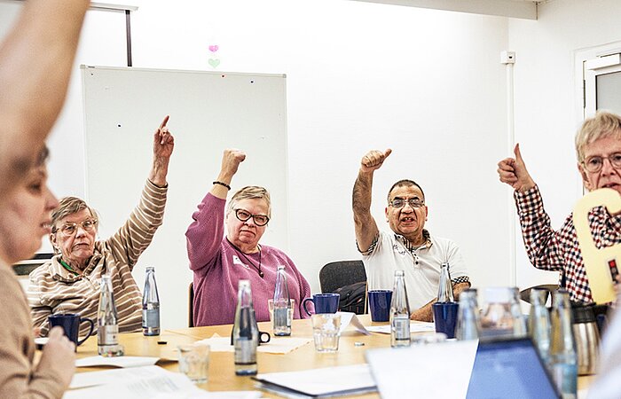 Mehrere Menschen sitzen an einem Tisch in einem Workshop und stimmen mit erhobenem Daumen über eine Frage ab.
