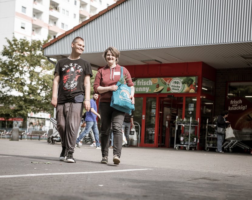 Zwei Menschen kommen lachend aus einem Supermarkt und tragen Einkaufstaschen. Im Hintergrund sind weitere Personen und Einkaufswagen zu sehen.