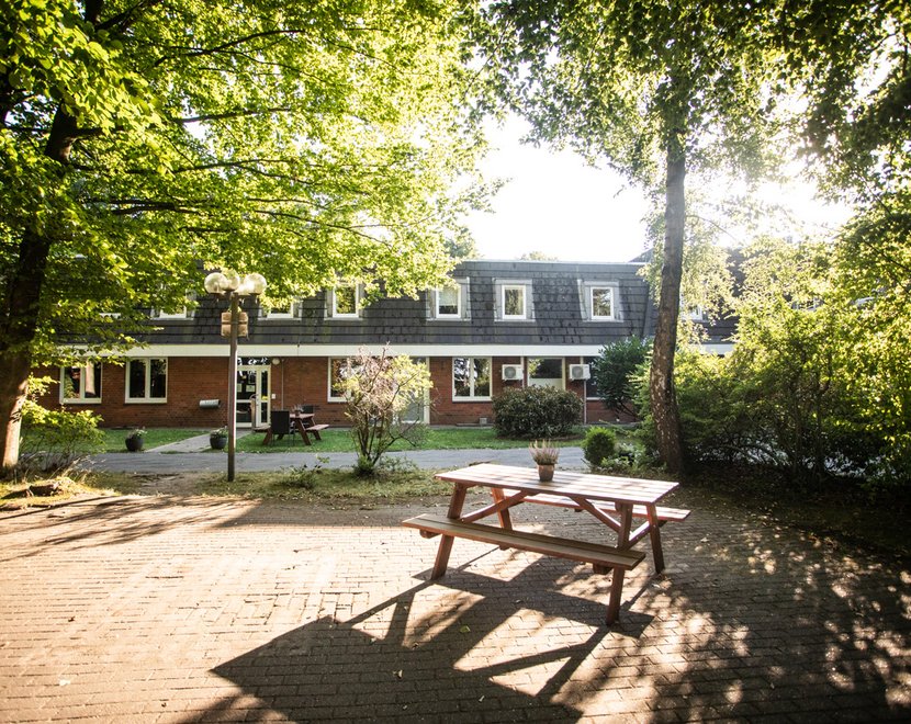 Haus Eckel von außen: Ein Backsteingebäude mit weißem Giebel liegt eingebettet in viel Grün. Im Vordergrund steht eine Sitzgruppe auf einem schattigen Platz unter Bäumen.