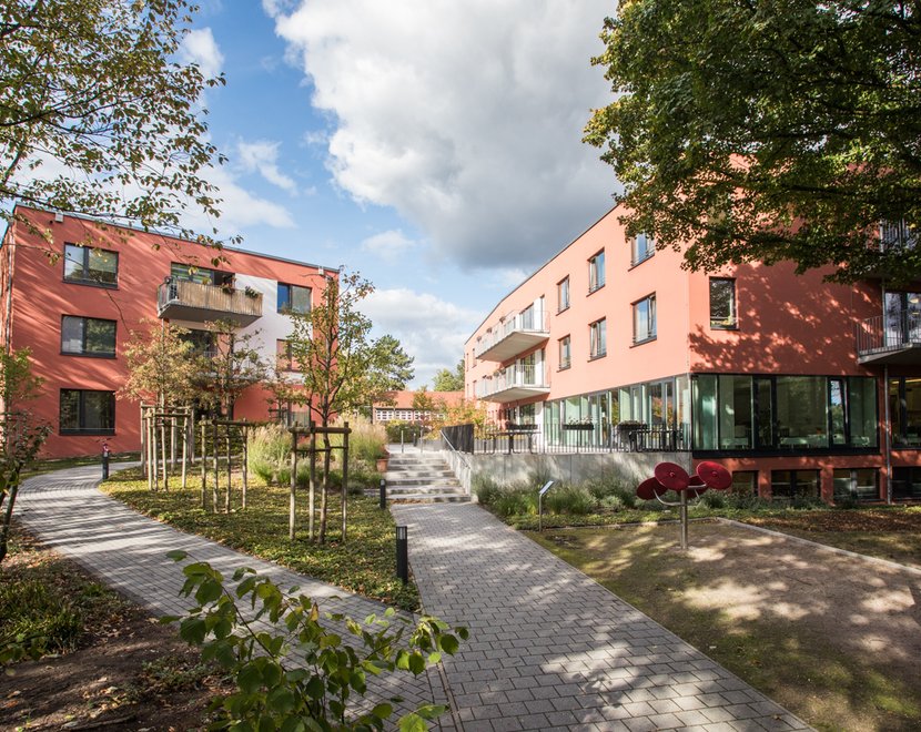 Außenansicht einer modernen, barrierefreien Wohnanlage mit zwei rot verklinkerten Gebäuden, breiten Wegen und viel Grün. Kleine Bäume und Sträucher säumen die Wege, die durch den begrünten Innenhof führen. Die Szene wirkt ruhig und einladend und zeigt ein Wohnangebot des Sozialkontors für Menschen mit Unterstützungsbedarf in einem inklusiven Umfeld.