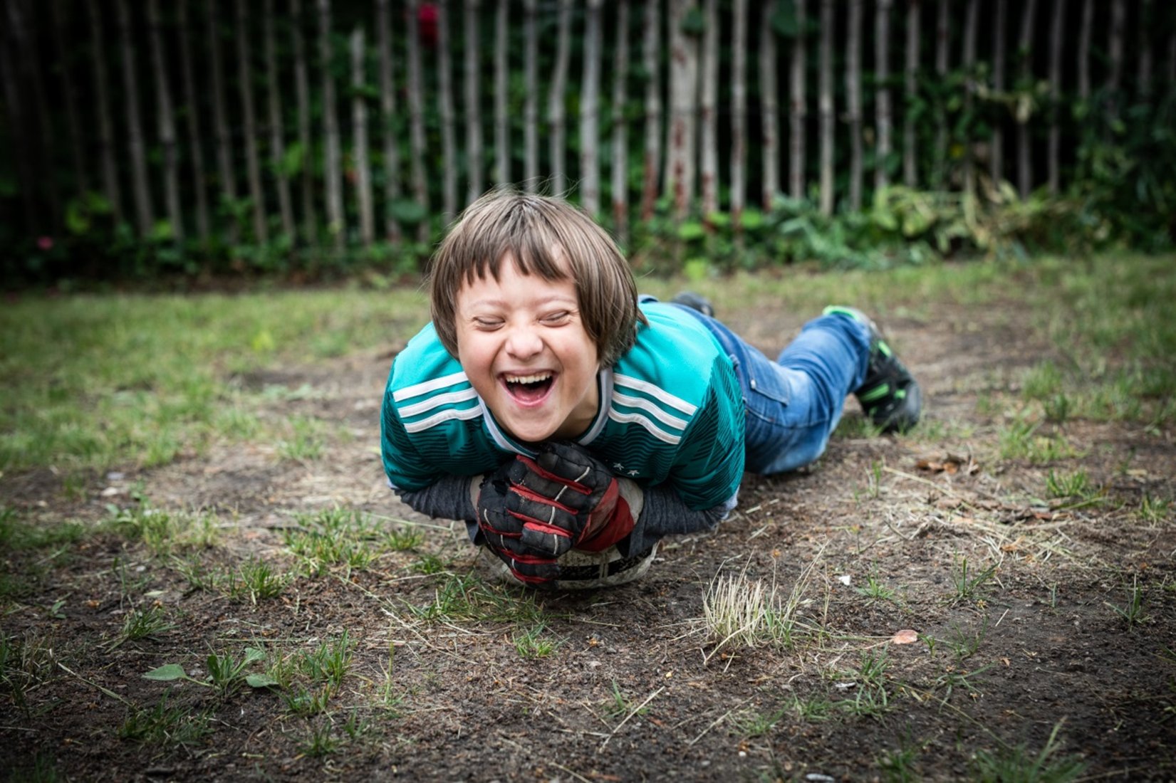 Ein lachender Junge in einem grünen Fußballtrikot liegt mit ausgebreiteten Armen bäuchlings auf dem Boden, während er einen Fußball hält. Im Hintergrund ist ein Holzzaun und etwas Grünfläche zu sehen.