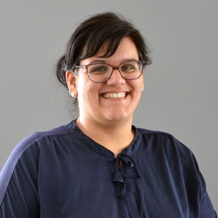 Sarah Bartsch, eine Frau mit dunklem, gelocktem Haar und Brille, lächelt freundlich in die Kamera. Sie trägt ein dunkelblaues Oberteil. Der Hintergrund ist neutral grau gestaltet.