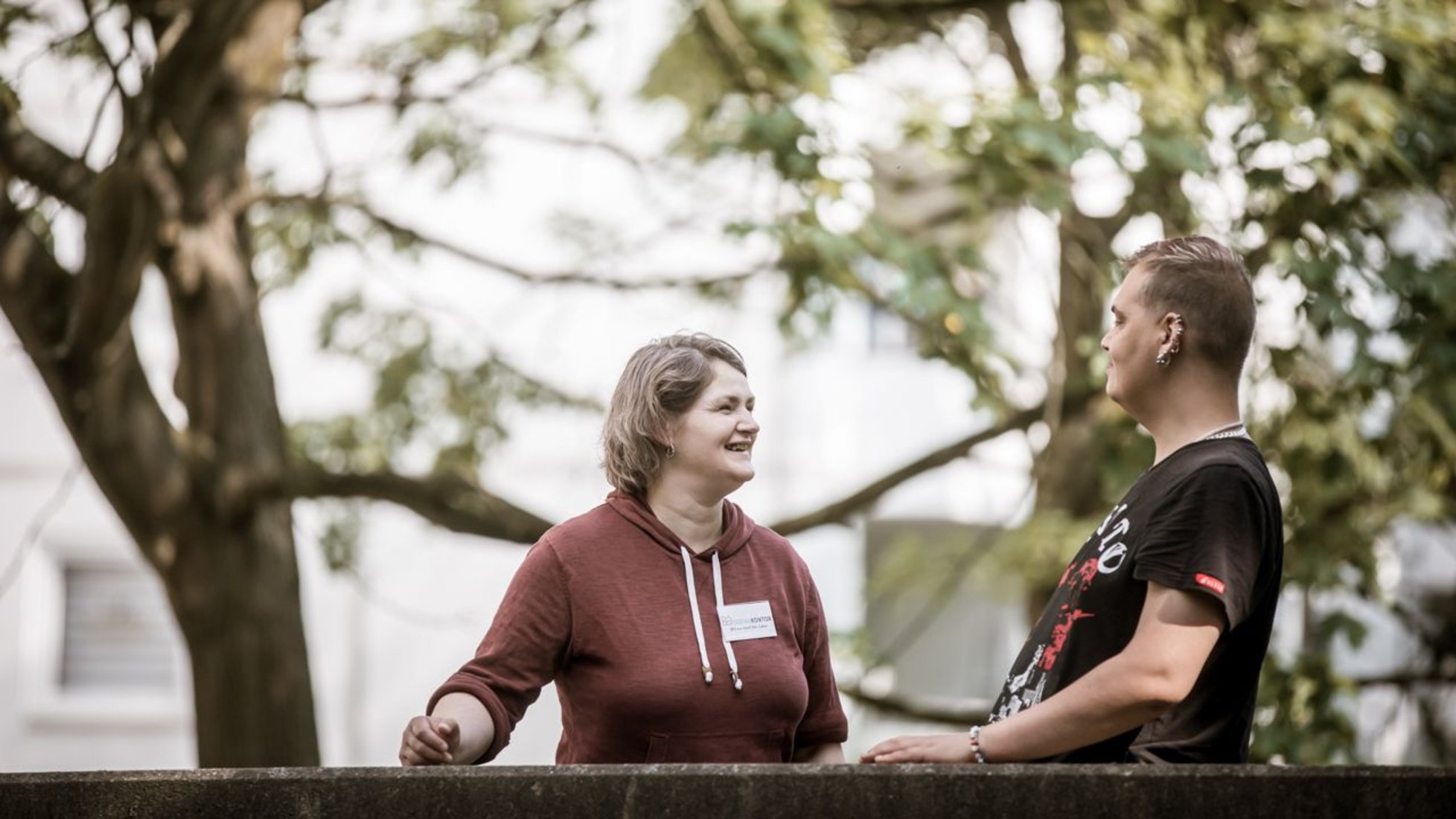 Zwei Personen stehen im Freien an einem Geländer und unterhalten sich angeregt und lachend. Im Hintergrund sind grüne Bäume und ein Gebäude zu sehen.