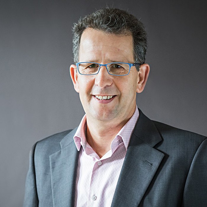 Gerd Müller, Geschäftsführung Finanzen und IT, ein Mann mit Brille und kurzem Haar, lächelt freundlich in die Kamera. Er trägt ein dunkles Jackett über einem rosa Hemd. Der Hintergrund ist dunkelgrau gestaltet.