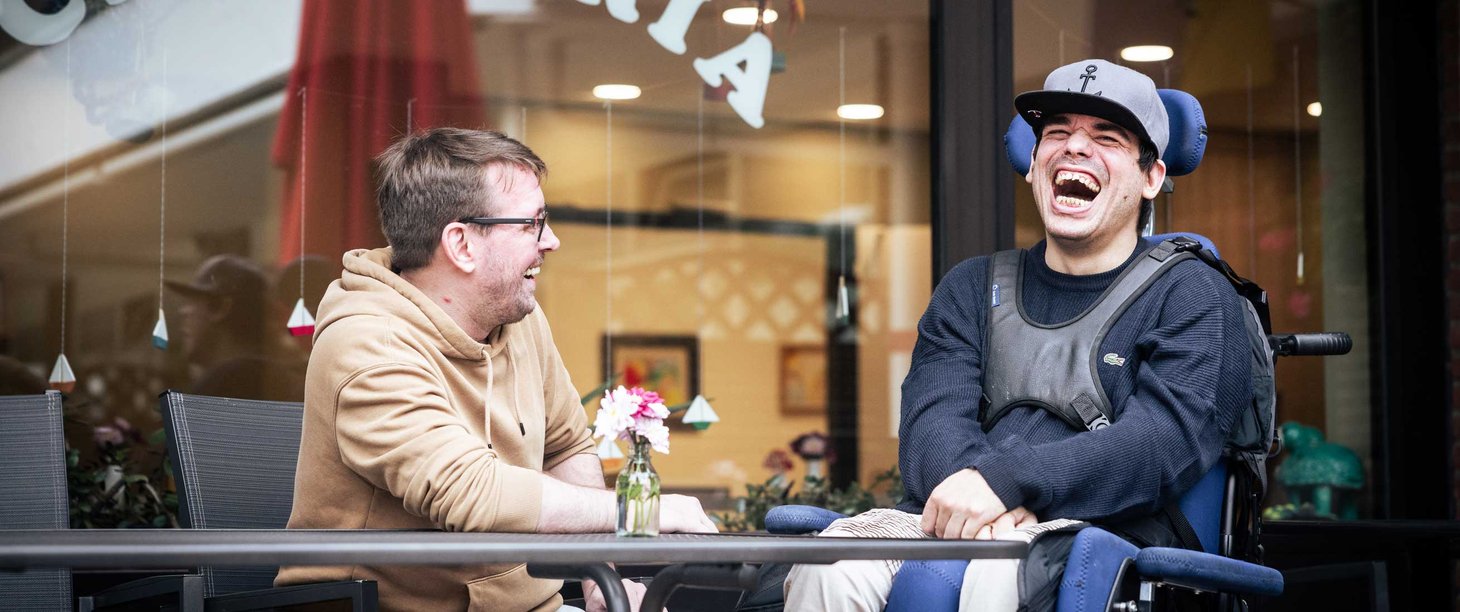Zwei Männer sitzen an einem Tisch vor einer Cafeteria und lachen. Der Mann rechts sitzt in einem Rollstuhl.