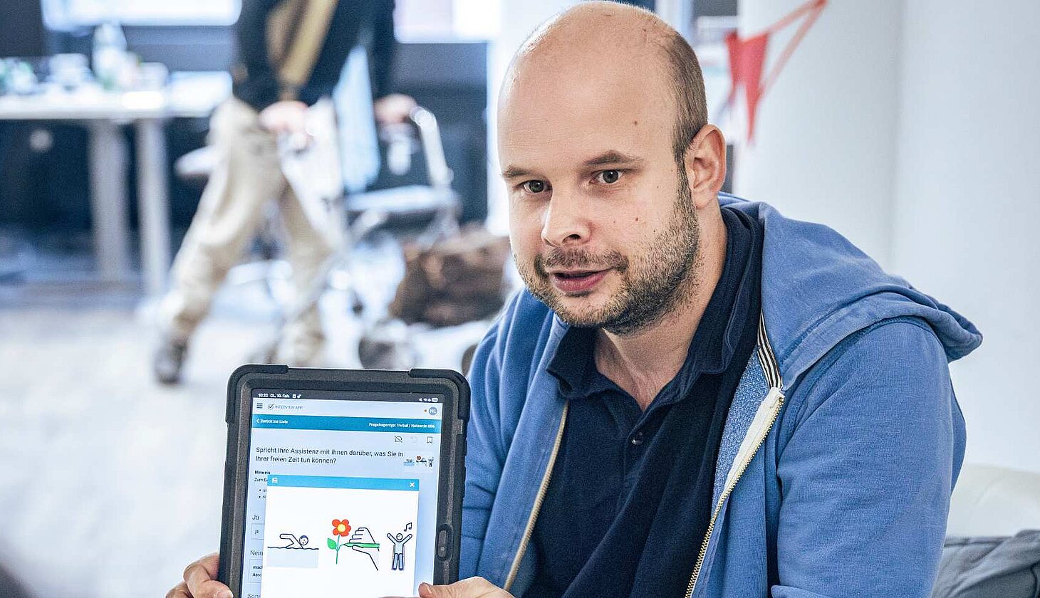 Niklas Luy zeigt ein Tablet mit Symbolen aus einer Befragungs-App. Die Bilder helfen dabei, Fragen zur Assistenz verständlicher zu machen.