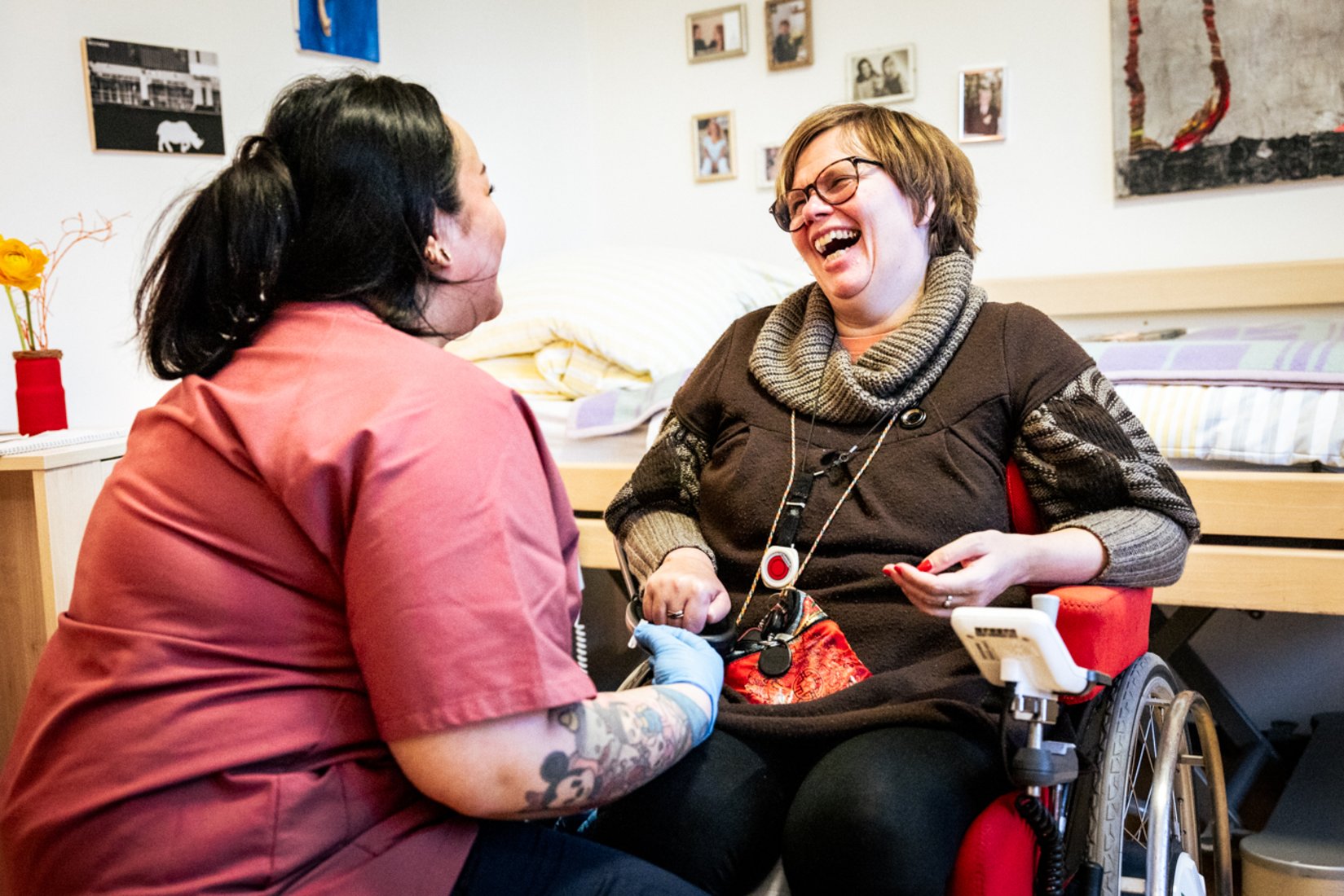 Eine Frau im Rollstuhl und eine Assistenzkraft in roter Arbeitskleidung sitzen einander zugewandt und lachen herzlich miteinander. Die Atmosphäre ist warm und vertraut. Im Hintergrund sind persönliche Gegenstände und Fotos zu sehen, die eine wohnliche Umgebung schaffen.