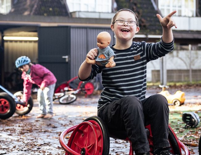 Förderschüler sitzt lachend auf einem Spielgerät im Freien und hält eine Puppe in der Hand, umgeben von anderen Kindern mit Fahrzeugen.