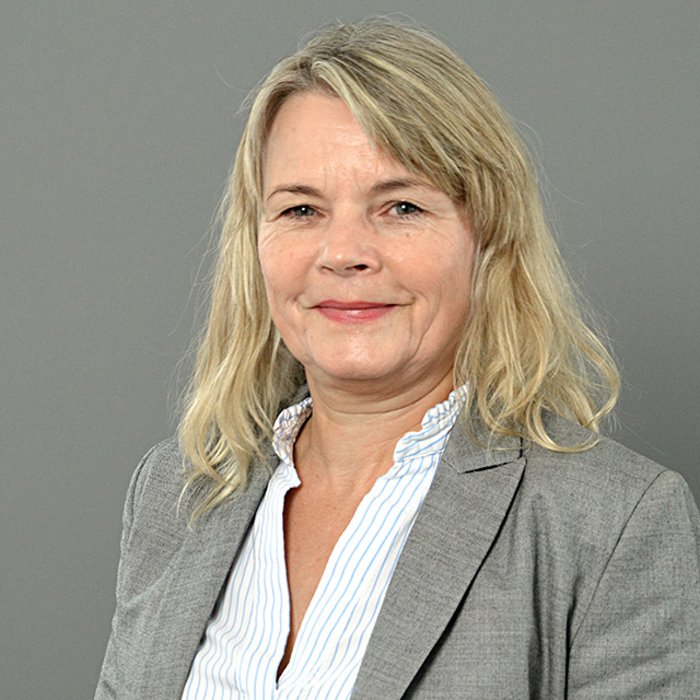 Nicola Pantelias, eine Frau mit lockigem, blondem Haar und Brille, lächelt freundlich in die Kamera. Sie trägt ein dunkles Oberteil. Der Hintergrund ist dunkelgrau gestaltet.