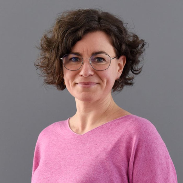 Eva-Maria Janulik, eine Frau mit lockigem, braunem Haar und Brille, trägt ein pinkfarbenes Oberteil. Sie blickt freundlich in die Kamera vor einem neutral grauen Hintergrund.